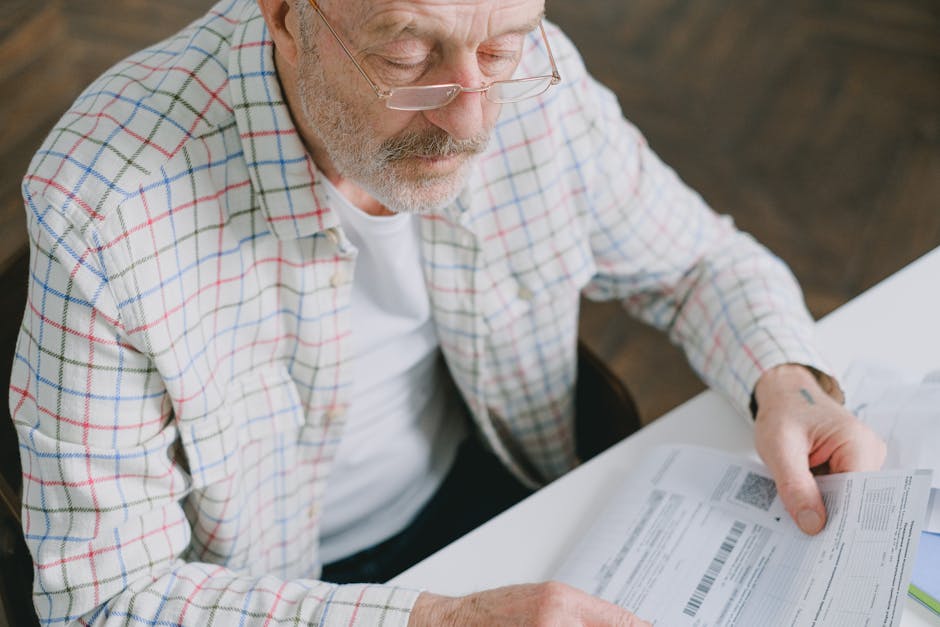 Elderly man in glasses carefully examines documents at home. Thoughtful and focused.