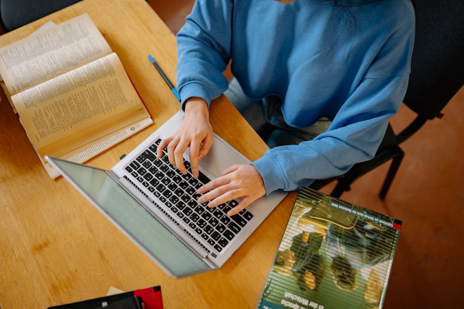 A student typing on a laptop surrounded by open books, focusing on research and school work.