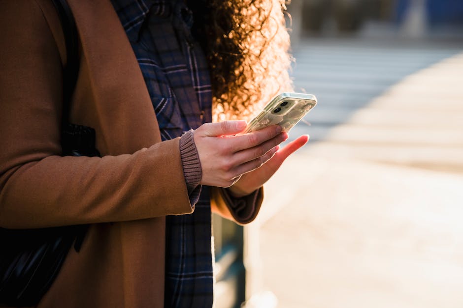 Crop anonymous female with curly hair in warm stylish coat using mobile phone on blurred background of urban street