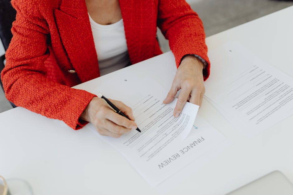 Close-up of a businesswoman in a red blazer signing finance documents at an office desk.