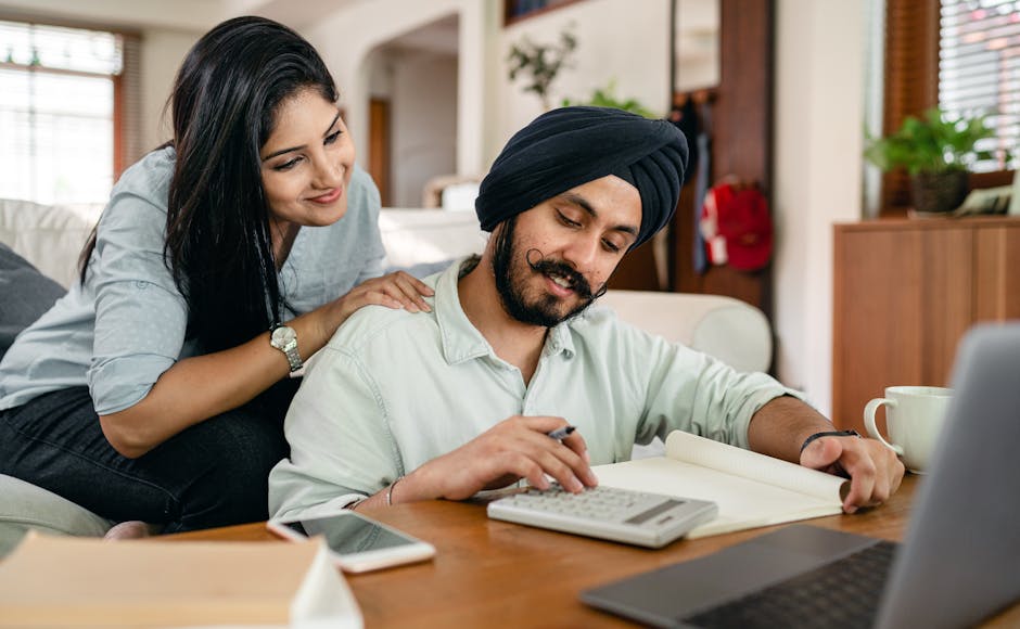 Concentrated ethnic male accountant using calculator with gadgets and books and notebook for remote work while ethnic wife touching shoulder of husband watching for numbers behind back sitting on sofa in living room