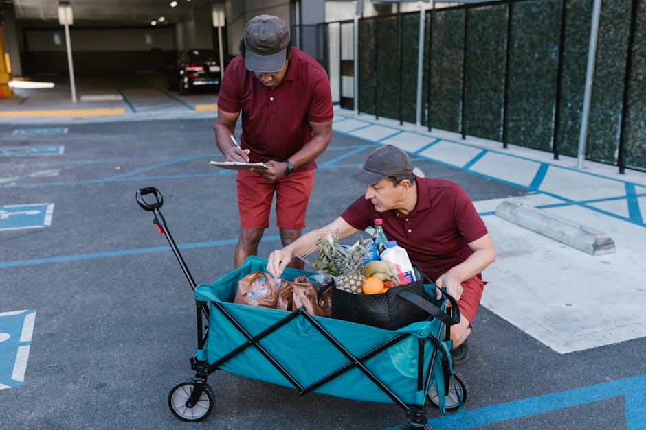Two men in red uniforms delivering groceries with a push cart in a parking area.