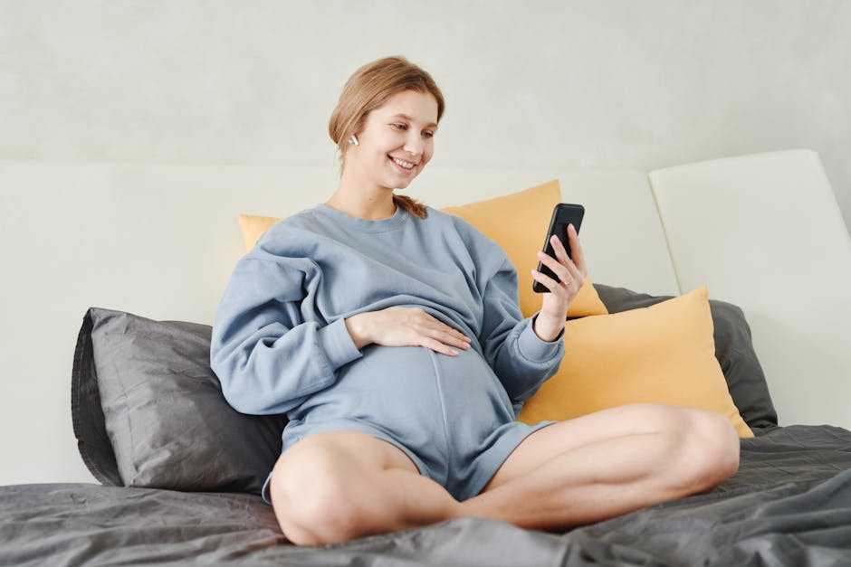 A pregnant woman sitting on bed smiling while using her smartphone indoors.