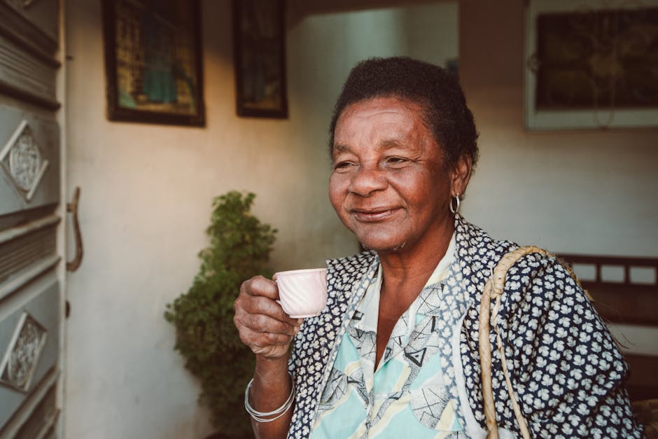 A senior black woman smiling warmly while holding a small coffee cup indoors.