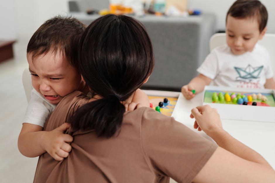 A mother comforts her crying child while another child plays nearby in a cozy home setting.