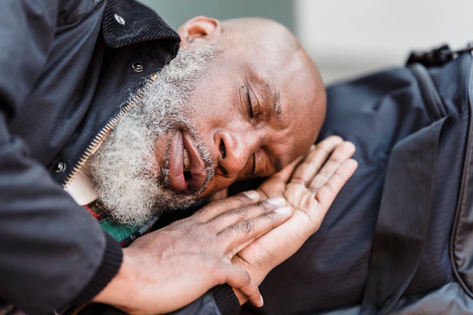 A bearded elderly man sleeps comfortably outdoors with a backpack as a pillow.