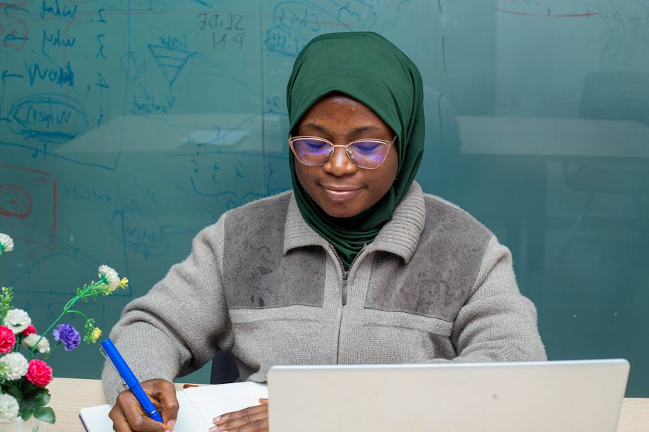 Woman in an office setting wearing a green headscarf, writing with laptop and notebook.