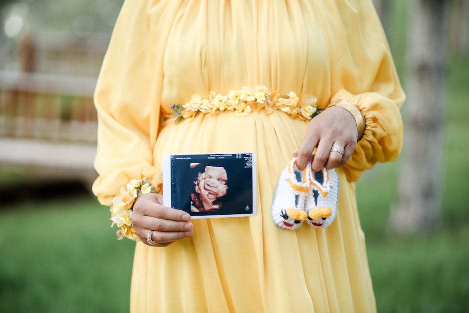 Pregnant woman in yellow dress holding ultrasound photo and baby shoes in a serene outdoor setting.