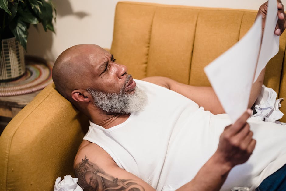 A bald man with tattoos looks at papers in disbelief while lying on a yellow sofa indoors.