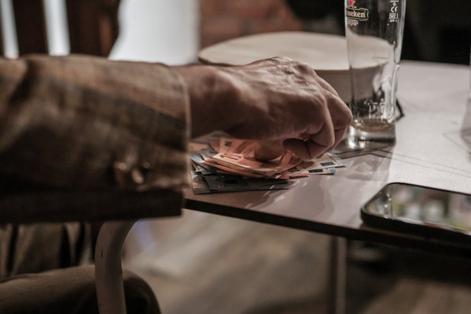 Hand counting euro banknotes on a table beside a phone and empty glass.