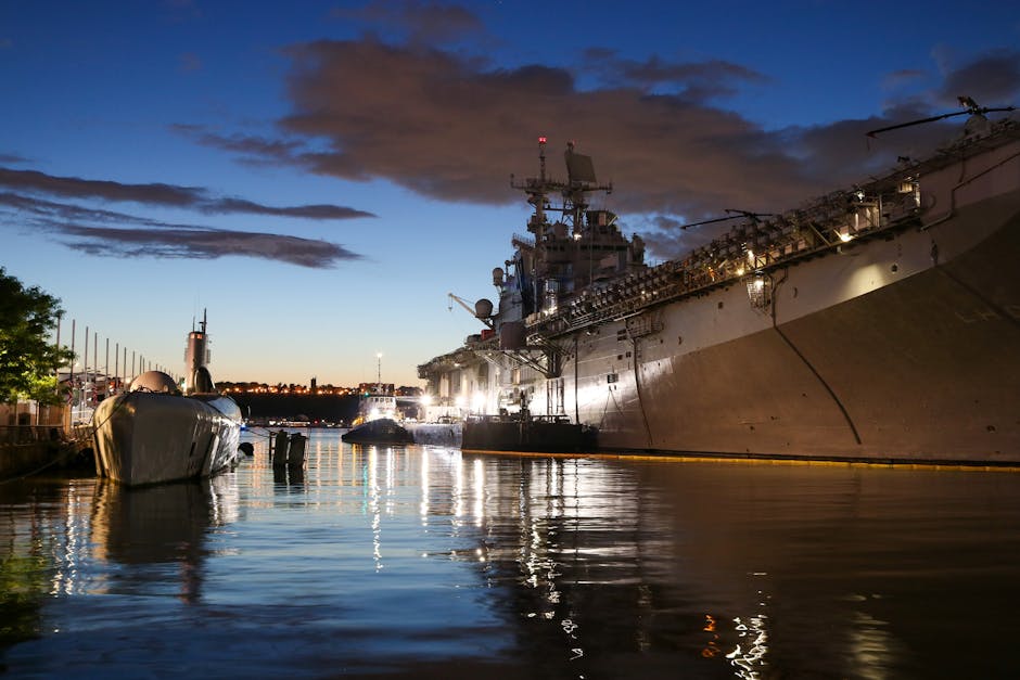 A naval warship docked in New York harbor at sunset, reflecting stunning lights on the water.