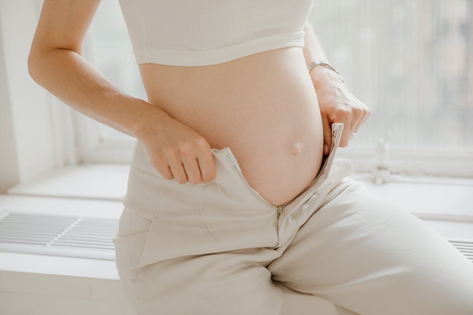 A close-up of a pregnant woman adjusting her pants, highlighting her belly in a sunlit room.