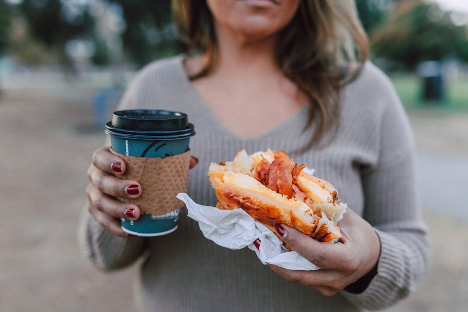 A woman holds a breakfast sandwich and a cup of coffee in an outdoor setting, capturing a casual morning moment.