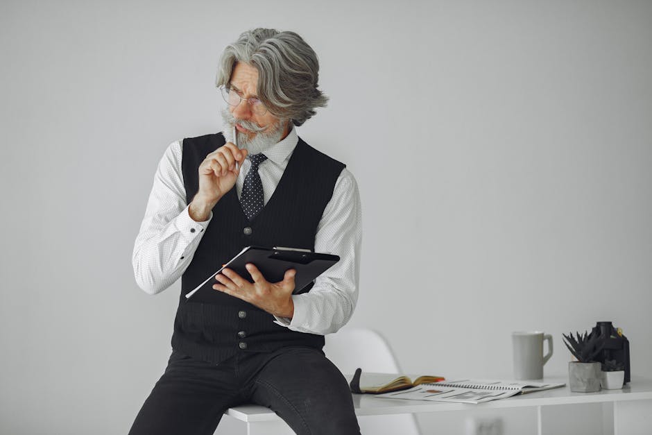 A thoughtful senior man with gray hair sits at a desk holding a tablet, reflecting in a modern office space.