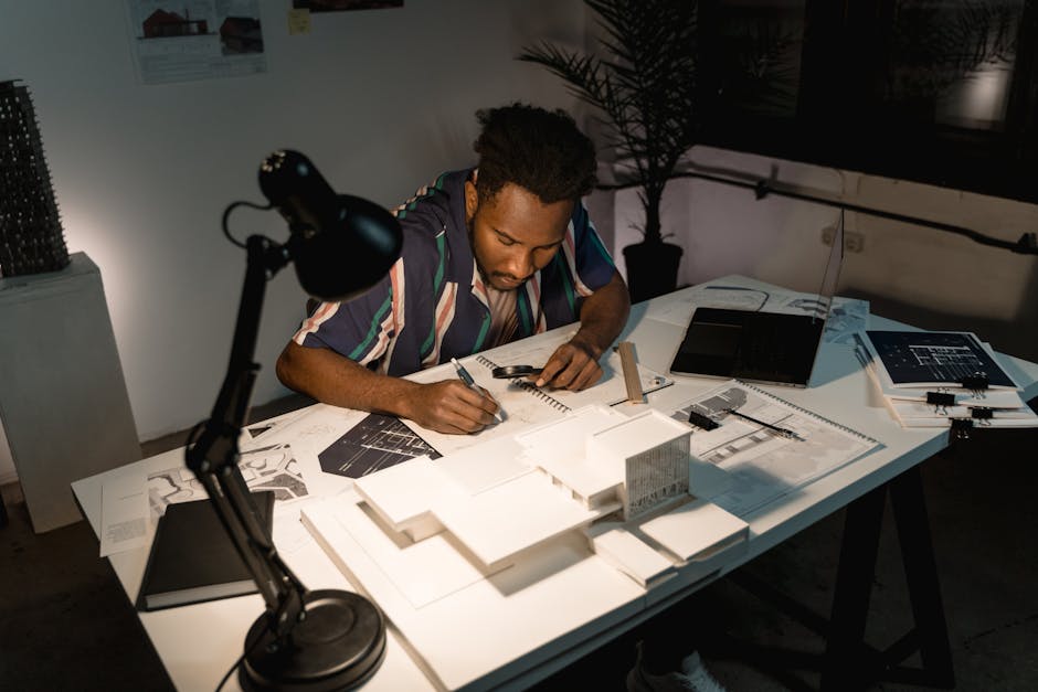 Focused architect working at desk, sketching designs with models and blueprints under desk lamp.