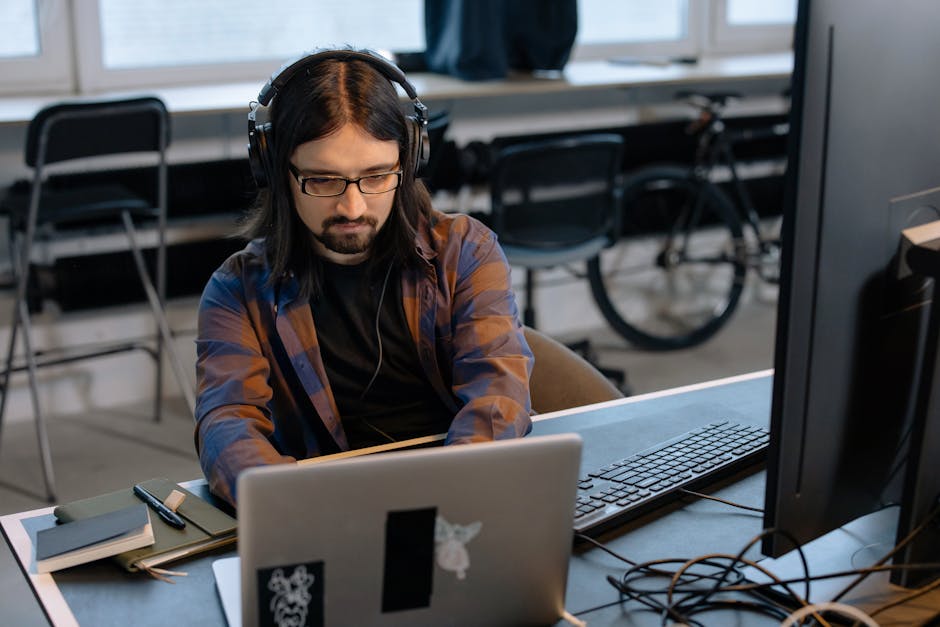 Bearded male developer in headphones coding on a laptop in a modern office setting.