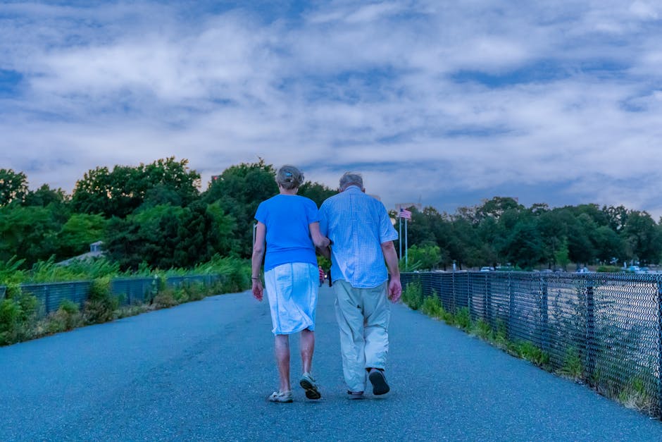 An elderly couple walking hand in hand on a peaceful outdoor pathway under a cloudy sky.