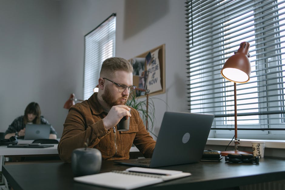 A bearded man drinking coffee while working on a laptop in a modern office space.