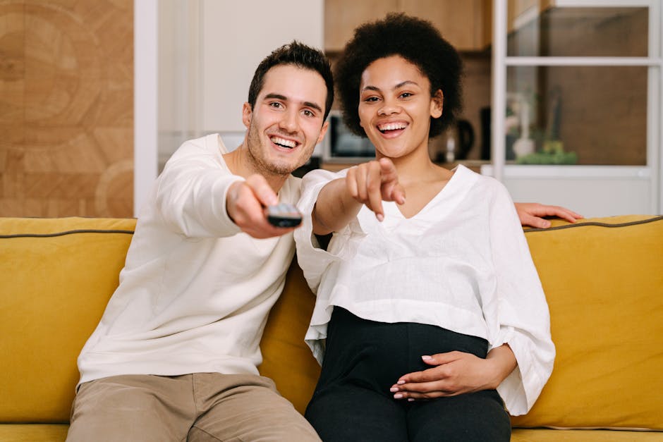 A smiling pregnant couple relaxes on a yellow sofa, enjoying movie time together.