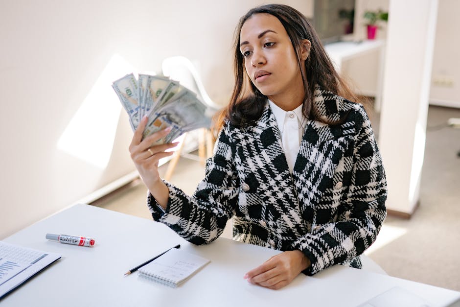 A young woman in a plaid coat counting US dollar bills at a desk in a modern office.