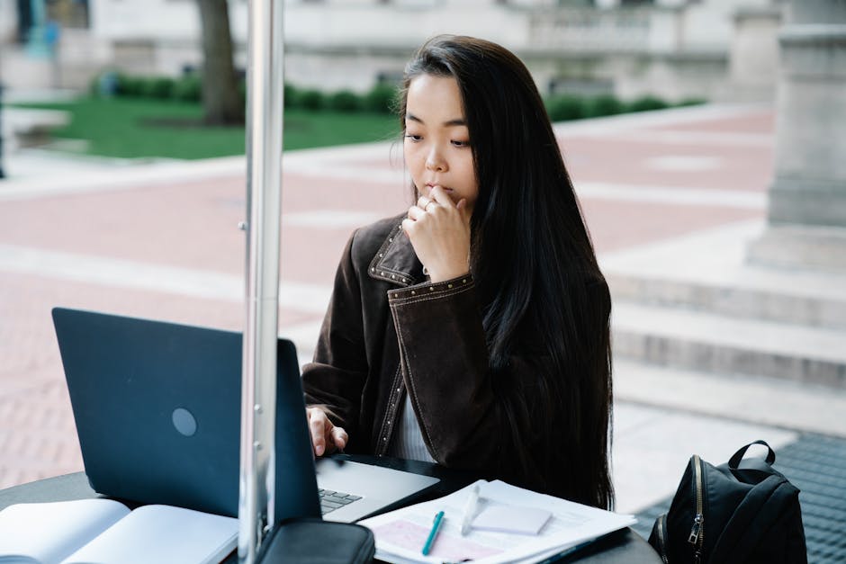 Young woman studying on laptop outside in a campus setting, concentrating on her work.