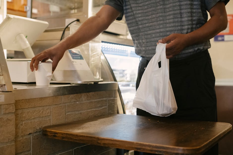 Unrecognizable male in casual clothes picking up order at counter with plastic bag and cup of hot drink in cafe
