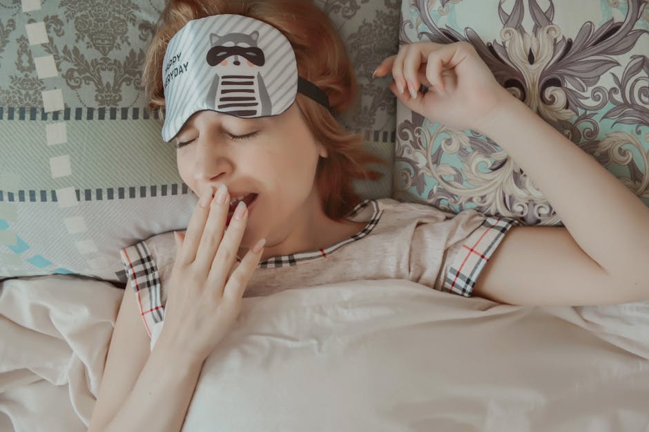 Top view of female with sleep mask wearing pajama covering mouth with hand while lying on bed with closed eyes under blanket during bedtime