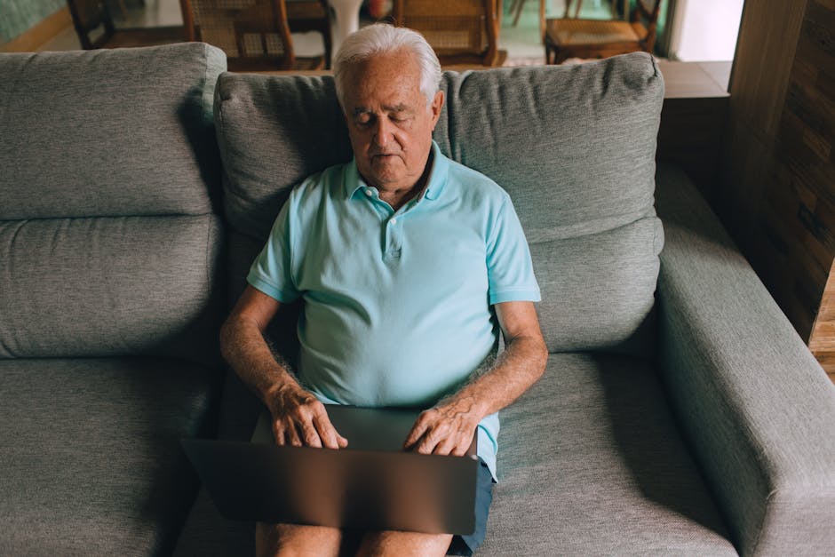 Elderly man working on a laptop while sitting on a cozy sofa in a home setting.