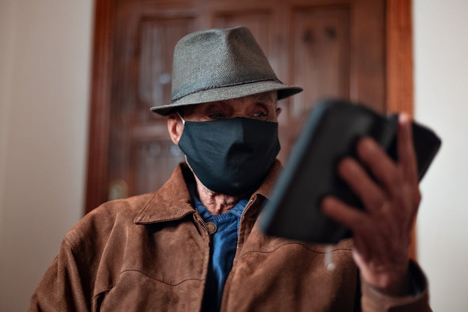 Senior male in hat and medical mask browsing smartphone while standing against blurred wooden door in light room in daytime