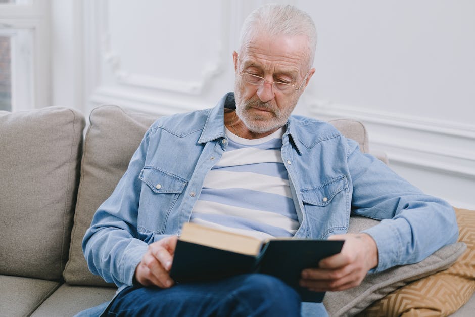 Elderly man with gray hair reading in a comfortable indoor setting, wearing casual clothes.