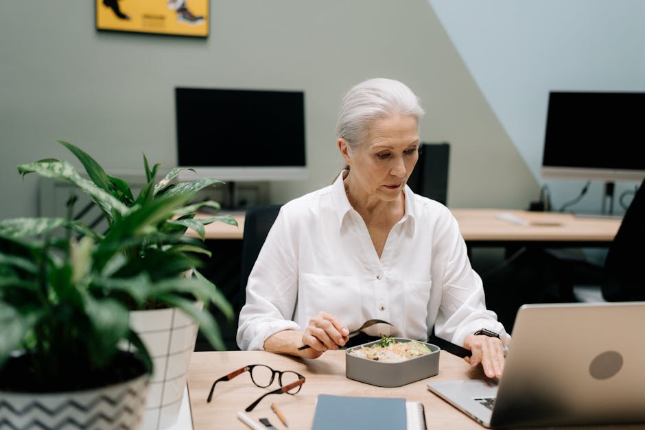 Elderly woman multitasking with lunch and work on laptop in an office setting.