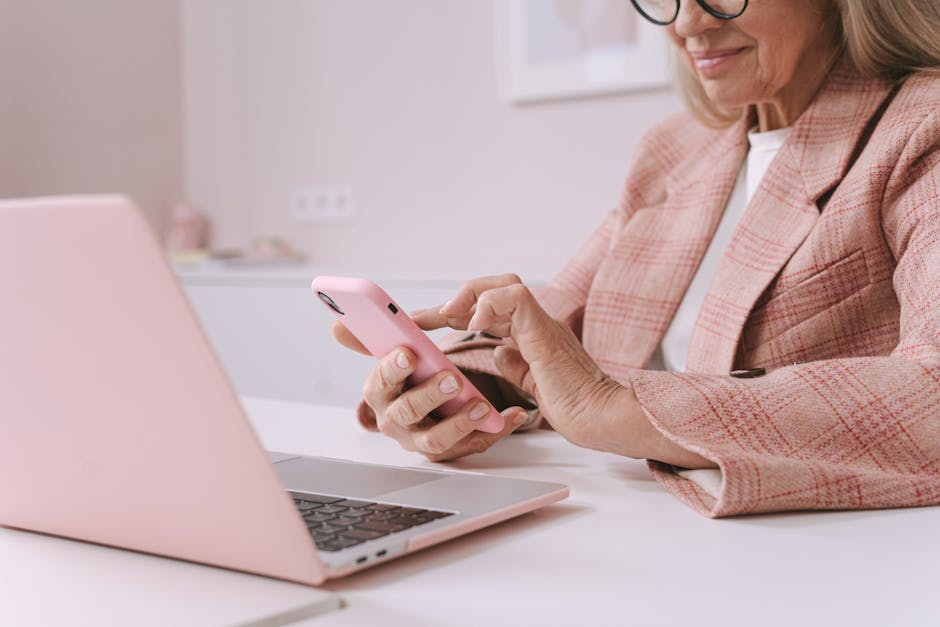 Elderly woman in pink plaid suit using a smartphone and laptop at a desk.