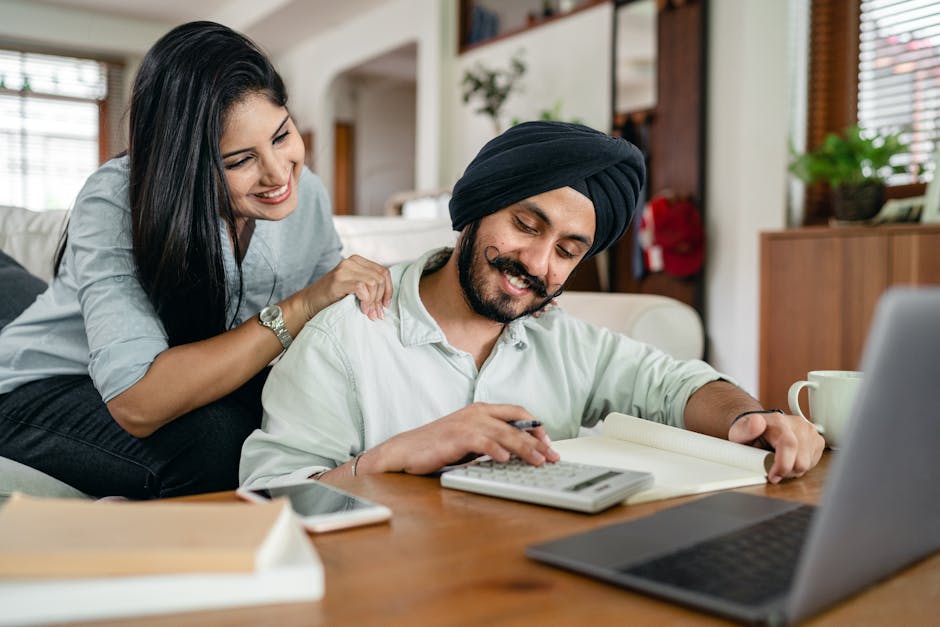 Positive ethnic male accountant using calculator and notebook and laptop and smartphone and books working at coffee table while ethnic wife sitting on sofa watching behind and touching husband back