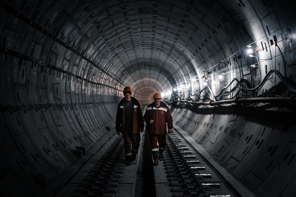 Two workers in a dark railway tunnel in Moscow, wearing uniforms and helmets, walking under lights.