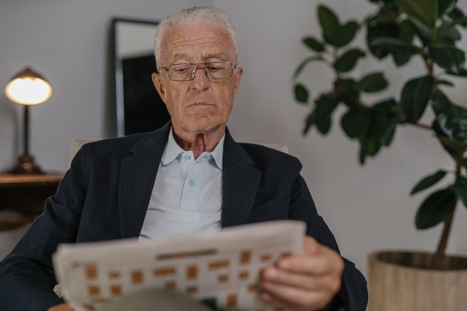 Senior man in suit jacket reading a newspaper indoors, near a plant.