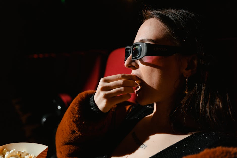A woman in 3D glasses enjoys popcorn while watching a movie in a theater setting.