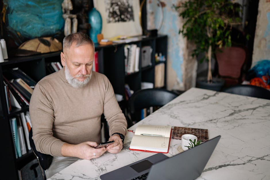 Elderly man using cellphone in a cozy home office setting with laptops and notebooks.