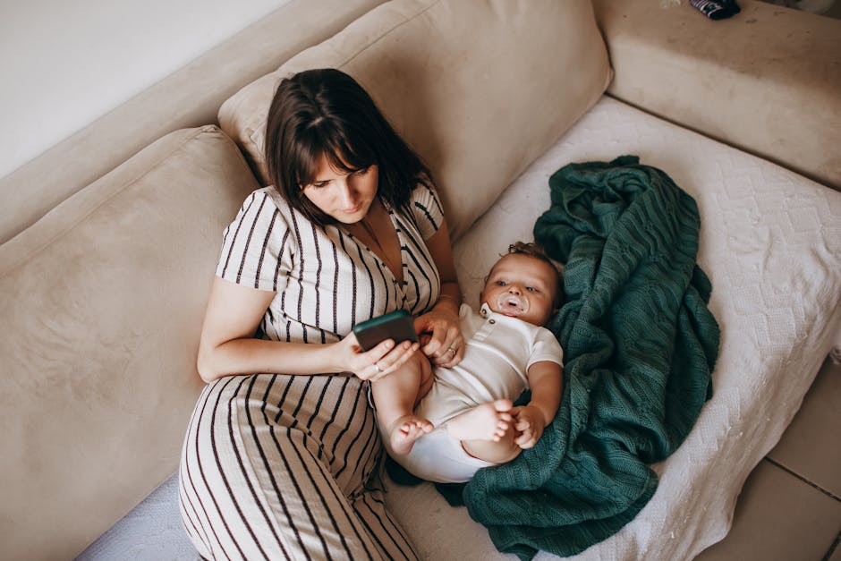 A mother and her baby relaxing on a beige couch browsing a smartphone indoors.
