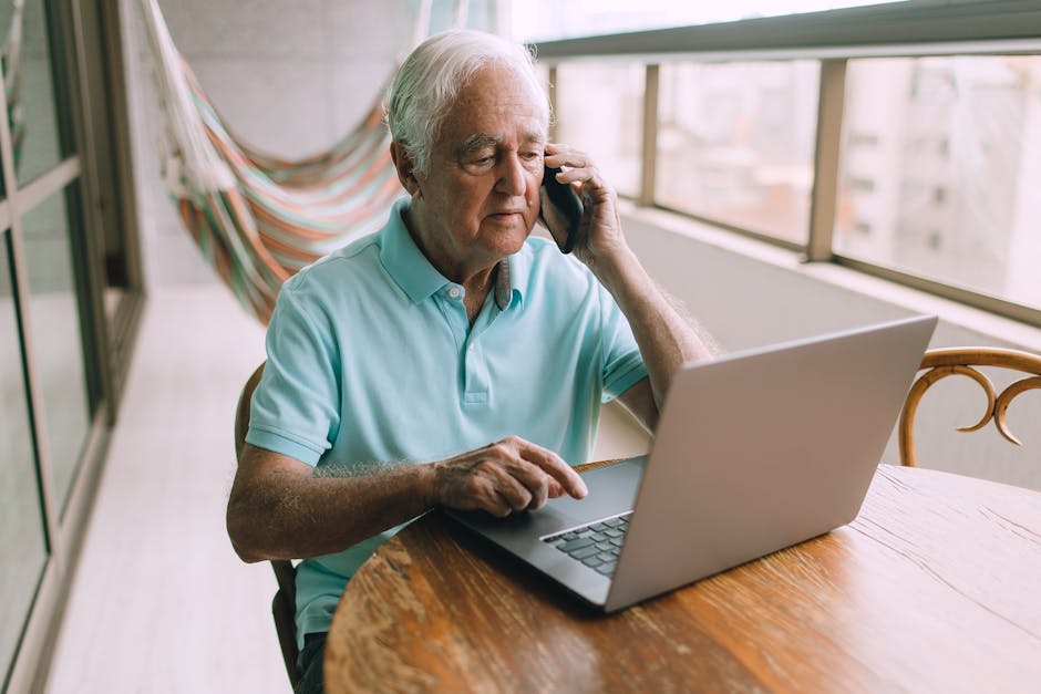 Elderly man multitasking on phone and laptop at home.