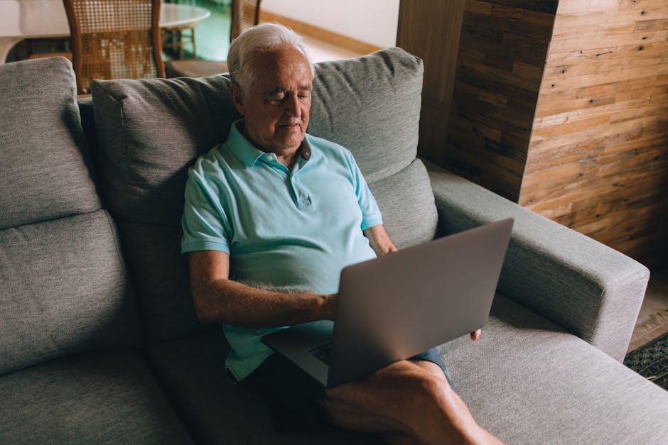 Senior man in blue polo shirt using a laptop while sitting on a cozy couch indoors.