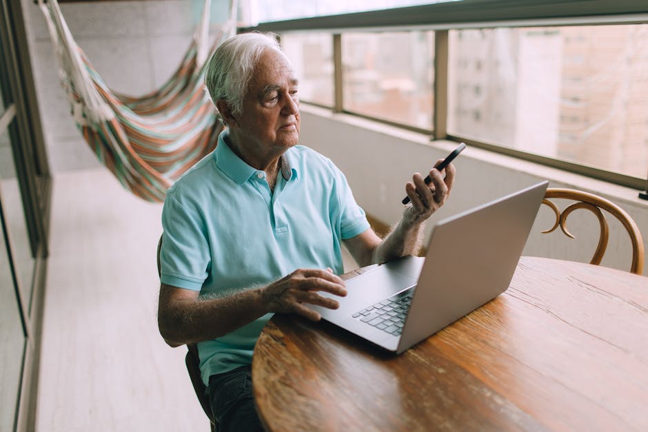 Elderly man multitasking with laptop and smartphone indoors, showcasing modern technology use.