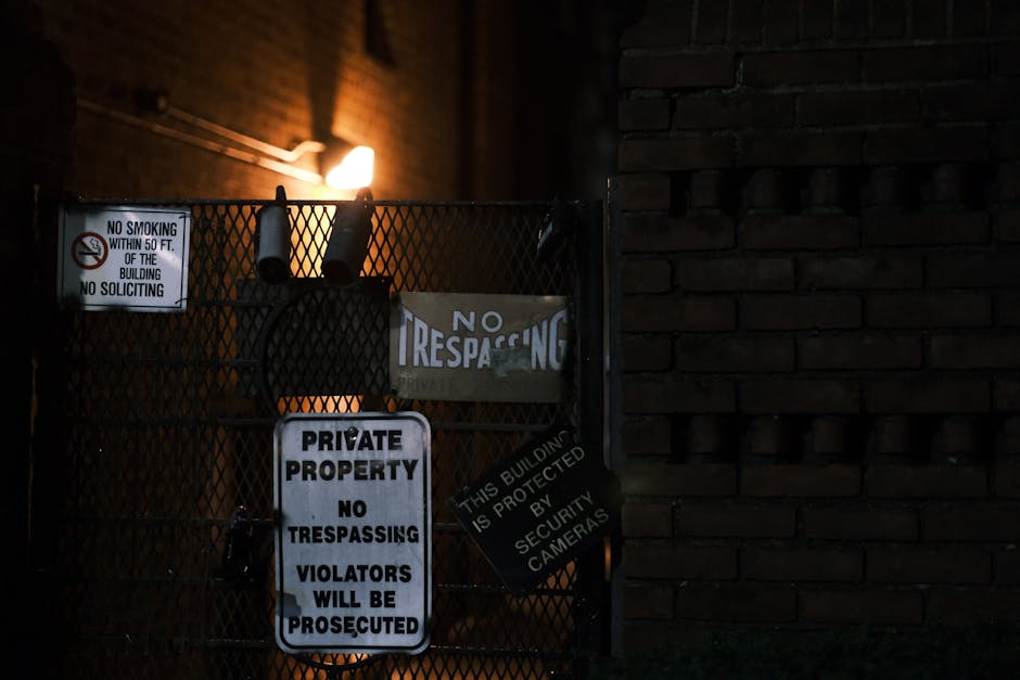Dark urban scene of a brick wall and gate with multiple warning signs.