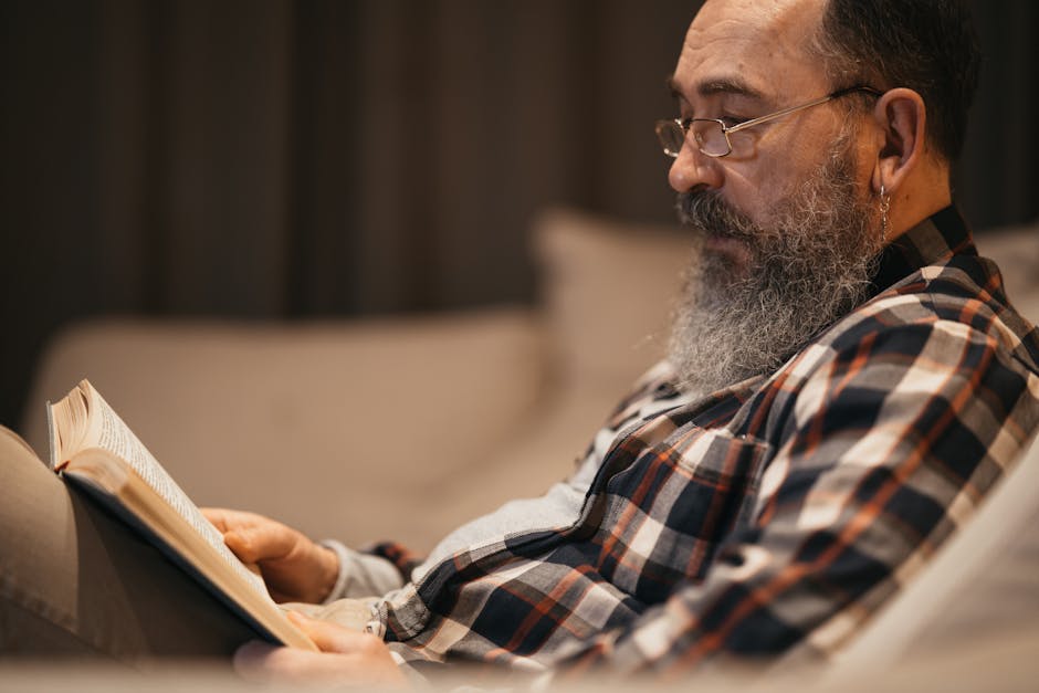 A senior man with a beard and glasses reading a book on a comfortable sofa indoors.