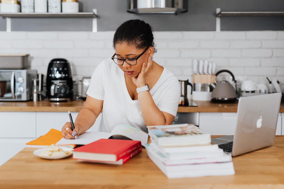 Woman studying at kitchen table surrounded by books and laptop, focused on writing.