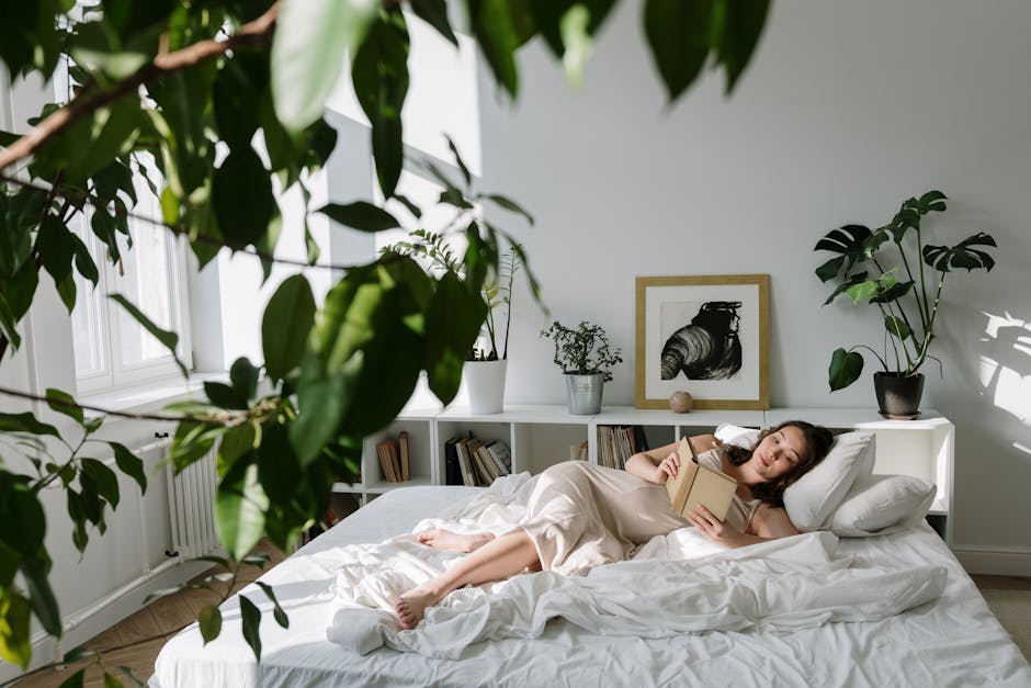 Pregnant woman reading a book in a cozy, plant-filled bedroom with sunlight streaming in.
