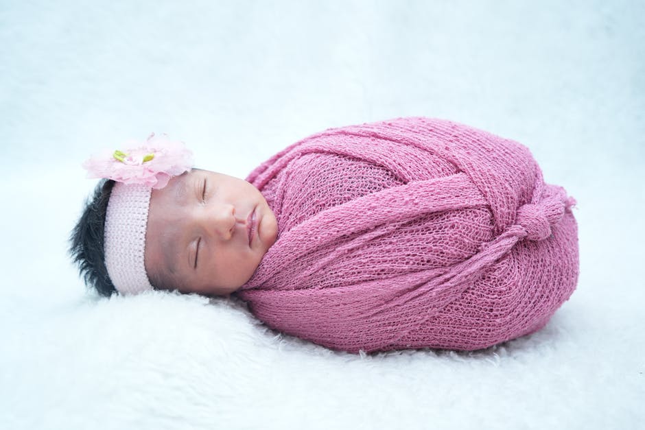 A peaceful newborn baby girl wrapped in a pink knitted blanket with a floral headband.