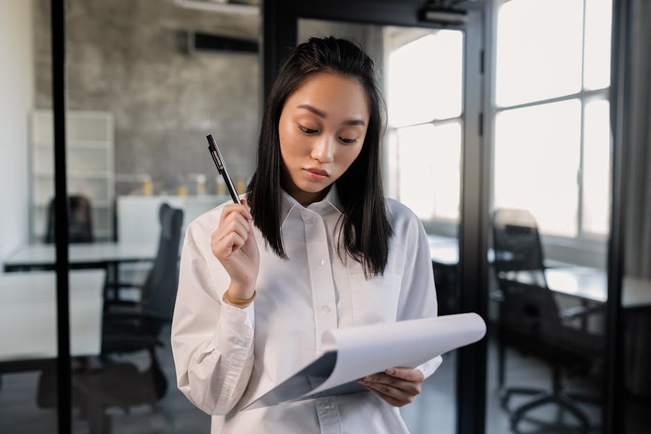 Confident Asian woman in office reviewing documents with pen.