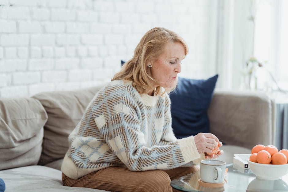 Side view of middle aged female with blond hair in stylish sweater sitting on soft couch and peeling fresh tangerines during breakfast at home