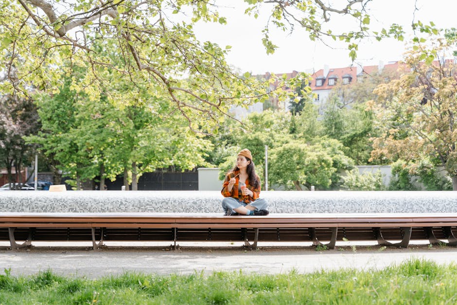 Asian woman sitting on a park bench, eating a snack on a sunny day surrounded by greenery.