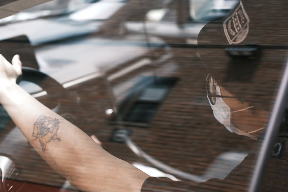 Person driving a car with a visible tattoo on arm seen through window reflection, wearing a cap and mask.